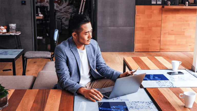 A man in a blue blazer sits at a wooden table, using a laptop and holding a smartphone, reflecting on an exciting partnership. The room has modern decor with a plant on the table and a coffee mug beside him. The atmosphere is calm and professional, reminiscent of an InMobi office setup.