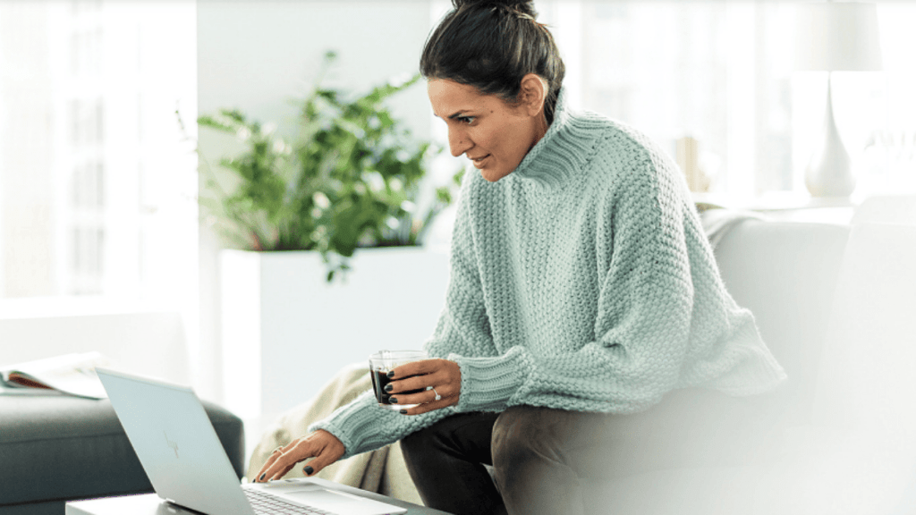 A person in a cozy sweater sits on a sofa, holding a small cup and using a laptop to browse Microsoft Advertising. The bright, airy environment features a potted plant in the background.