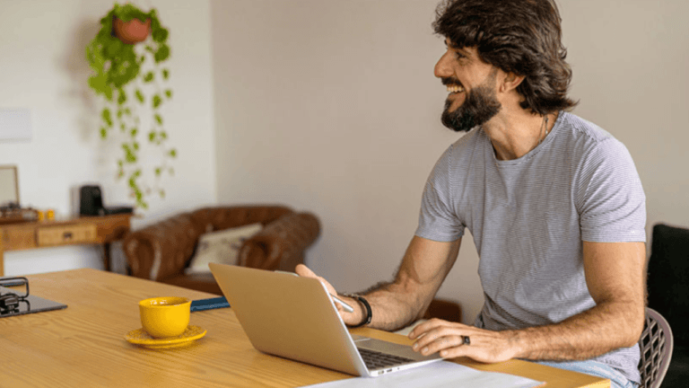 A bearded man is seated at a wooden table, smiling as he uses his laptop featuring Microsoft tools. Beside him, a yellow mug complements the cozy room adorned with a brown couch and a hanging plant, enhancing his globally inspired workspace.