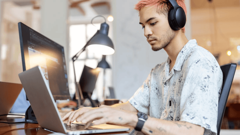 A person with pink hair and headphones sits at a desk, working intently on a laptop. Nearby, a large monitor displays their vision for the future. A desk lamp casts light, while sunlight filters through the windows, illuminating office decor in this Microsoft-inspired workspace.