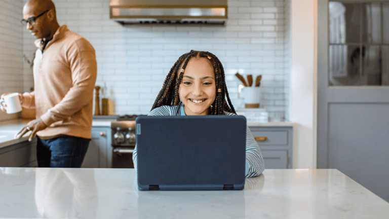 A young girl with braided hair smiles while using a Microsoft laptop on a kitchen island. In the background, a man holds a mug, standing by the stove. The kitchen has a modern design with white subway tile and gray cabinets, blending seamlessly into this future-ready setting.