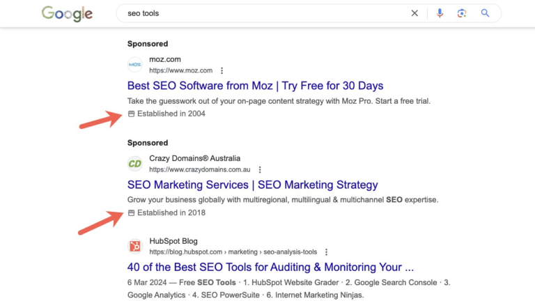 Screenshot of a Google search for "SEO tools" showcasing desktop search ads from Moz, Crazy Domains Australia, and HubSpot Blog. Each ad includes a brief description and highlights the company establishment year, marked with a red arrow.