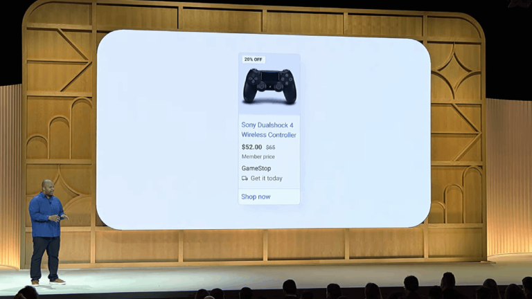 A speaker is on stage presenting a large screen displaying a Sony Dualshock 4 Wireless Controller advertisement. Highlighting a 20% discount, it’s priced at $52.00 for members under Loyalty Promotions, available at GameStop with an option to "Shop now.