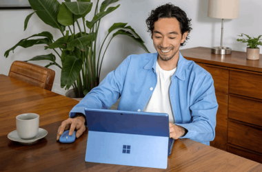 A person sits at a wooden table using a blue tablet and mouse. They're smiling, wearing a light blue shirt. A cup of coffee and a potted plant are nearby, with a wooden cabinet and lamp in the background.