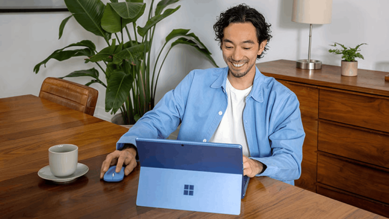A person sits at a wooden table using a blue tablet and mouse. They're smiling, wearing a light blue shirt. A cup of coffee and a potted plant are nearby, with a wooden cabinet and lamp in the background.