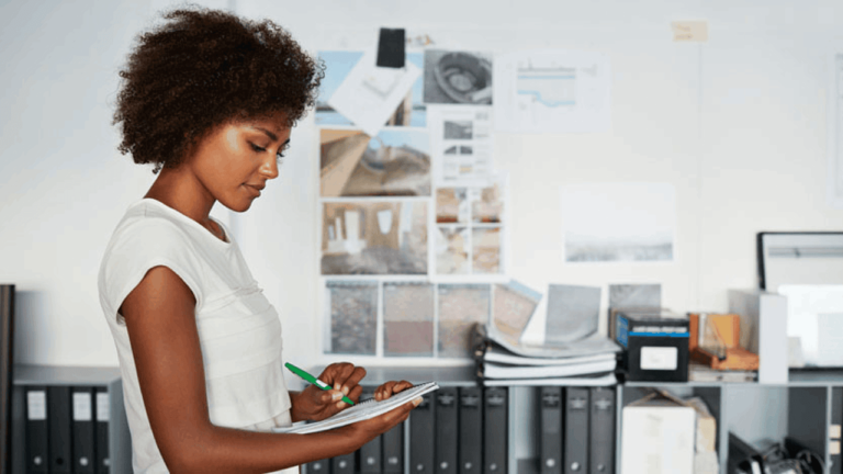 A woman with curly hair stands in an office, writing on a clipboard. Behind her, a wall displays several photographs and documents. Shelves with binders and files are visible in the background. She appears focused on her task.