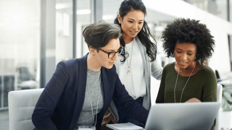 Three women are gathered around a laptop in an office setting. One is sitting and typing, while the other two stand beside her, all focused on the screen. Bright natural light fills the room, creating a collaborative atmosphere.