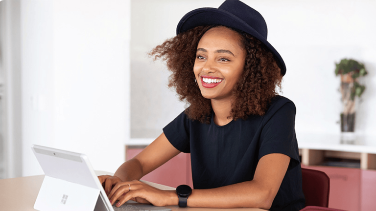 A person with curly hair is wearing a black hat and shirt, sitting at a desk and smiling while using a laptop, possibly optimizing campaigns through the Microsoft Advertising Platform. The background features a minimalist, modern interior with soft lighting.