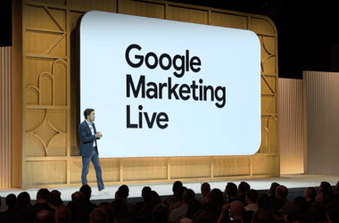 A speaker in a suit stands on stage, speaking in front of a large screen displaying the text "Google Marketing Live." The event has wooden paneling and a large audience is seated in front of the stage.