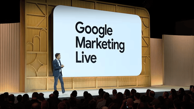 A speaker in a suit stands on stage, speaking in front of a large screen displaying the text "Google Marketing Live." The event has wooden paneling and a large audience is seated in front of the stage.