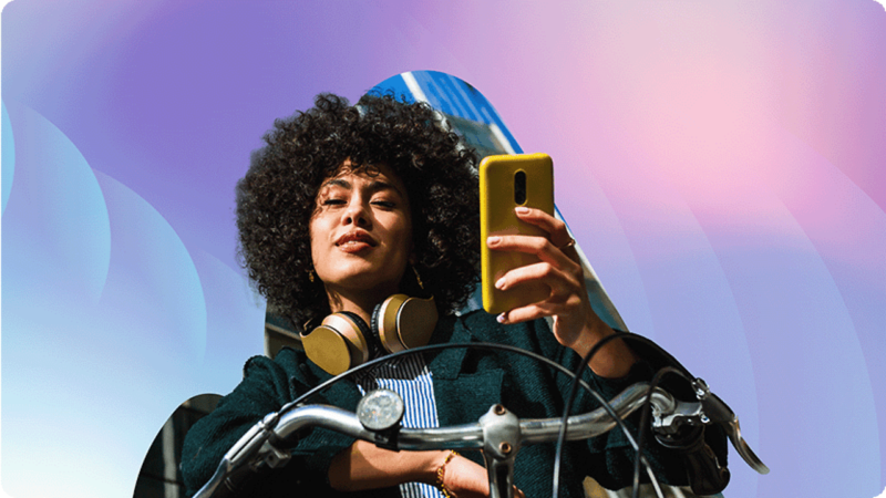 A woman with curly hair and headphones around her neck takes a personalized selfie with a smartphone while gripping bicycle handlebars. The backdrop showcases abstract, colorful shapes in blue and pink hues.