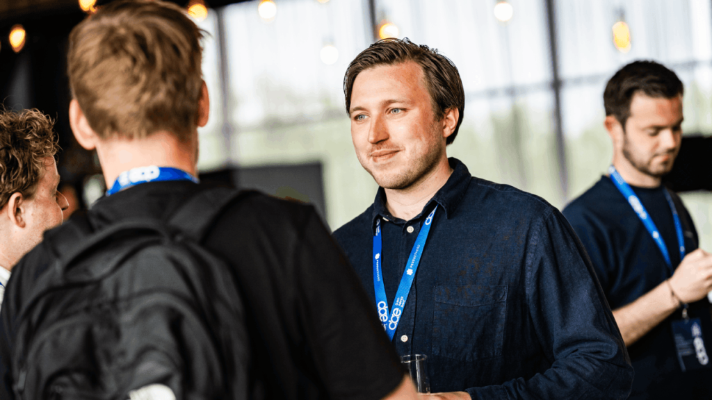 A group of people are conversing at an event. A man, holding a drink, is smiling and wearing a blue lanyard—perhaps discussing PPC advertising trends. Another person with a backpack is facing him. The blurred background with lights and windows adds to the lively atmosphere.