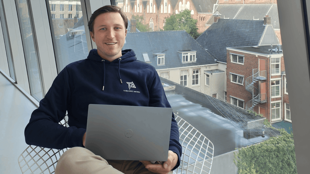 A person wearing a dark blue hoodie sits in a wire chair by a large window, holding a laptop. Behind them, an urban view with buildings and greenery is visible. They are smiling at the camera, preparing for an interview about PPC advertising strategies.