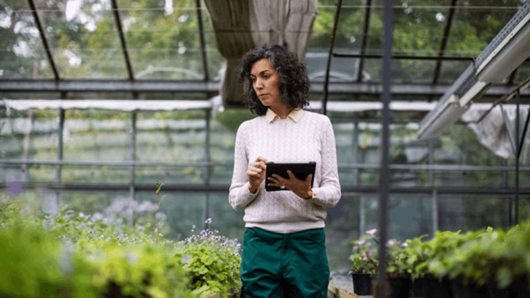 A woman with curly hair wearing a light sweater and green pants holds a tablet. She stands in a greenhouse surrounded by plants, appearing focused. The background shows a structure of glass and metal beams with greenery outside.