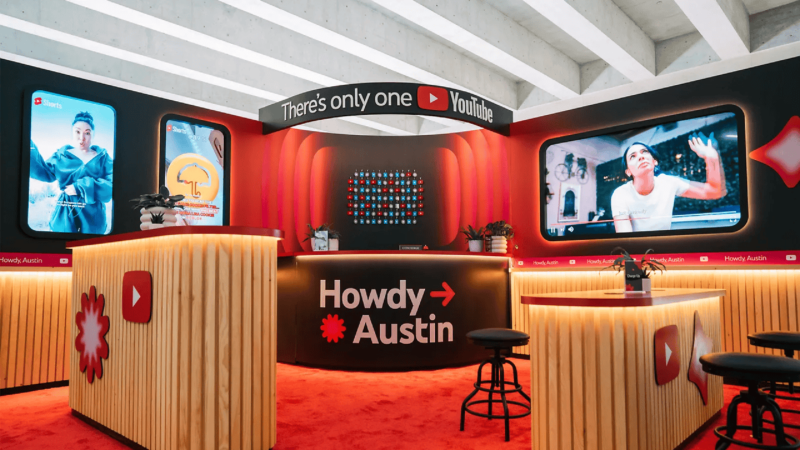 Brightly lit YouTube-themed booth with a "Howdy Austin" sign, featuring large digital screens, a red and wooden counter, and stools. Decorated with YouTube logos and red accents, this inviting space showcases new ways to work with creators through YouTube BrandConnect.