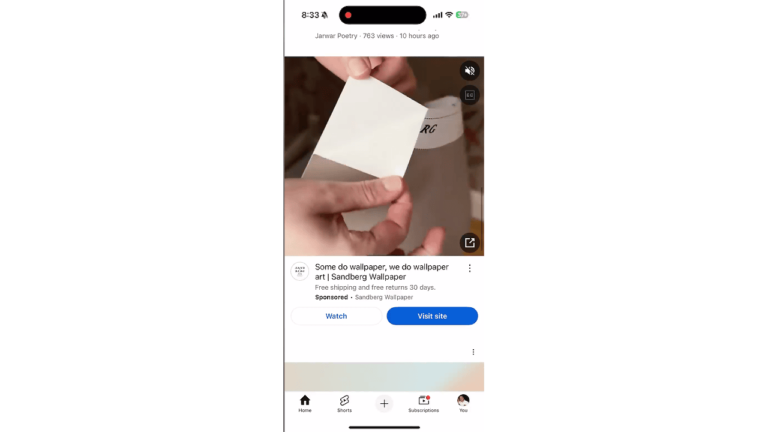 A person is applying adhesive wallpaper from the trending brand Sandberg Wallpaper onto a wall. The phone display shows a YouTube Ad with “Watch” and “Visit site” buttons, indicating it's a sponsored promotion.