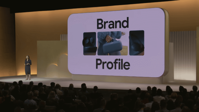 A woman presents on stage during GML 2025. Behind her, a large screen displays the words "Brand Profile" and three images of hands interacting with blue products, highlighting the importance of identity in search.