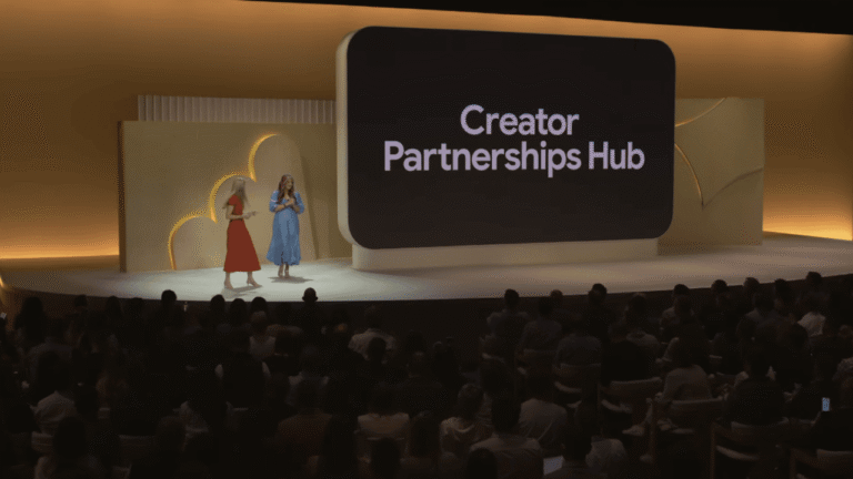Two women stand on a stage in front of a large screen displaying the words "Creator Partnerships Hub," addressing an audience in a dimly lit, modern event space.