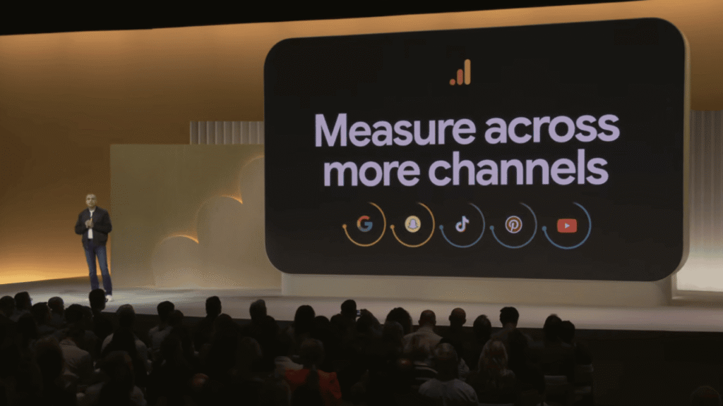 A speaker presents on stage beside a large screen displaying “Measure across more channels” with icons for Google, Meta, TikTok, Amazon, and YouTube—highlighting the importance of Cross-Channel Attribution for smarter budgeting decisions.