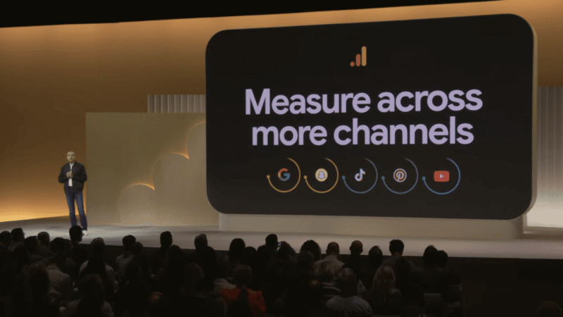 A speaker presents on stage beside a large screen displaying “Measure across more channels” with icons for Google, Meta, TikTok, Amazon, and YouTube—highlighting the importance of Cross-Channel Attribution for smarter budgeting decisions.