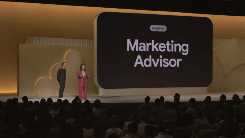 Two presenters stand on stage in front of a large screen displaying "Google Ads Marketing Advisor," discussing GML 2025 and real-time AI help with an audience in a dimly lit auditorium accented by yellow and gold decor.