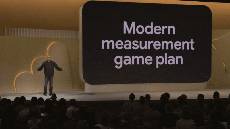 A speaker stands on stage in front of a large screen displaying the words "Modern measurement game plan" as an audience watches.