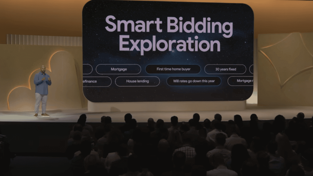 A speaker stands on stage before an audience, presenting a large screen that reads “Smart Bidding Exploration” and displays related keywords like “Mortgage,” “First time home buyer,” and highlights AI Max for Search Campaigns.