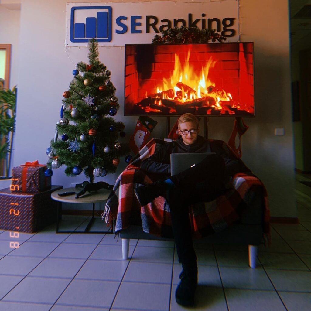 A person sits on a couch with a laptop in a cozy office decorated for Christmas, featuring a tree and stockings. A digital fireplace glows on the TV under the SE Ranking sign, while PPC Reporting tools provide valuable AI Insights.