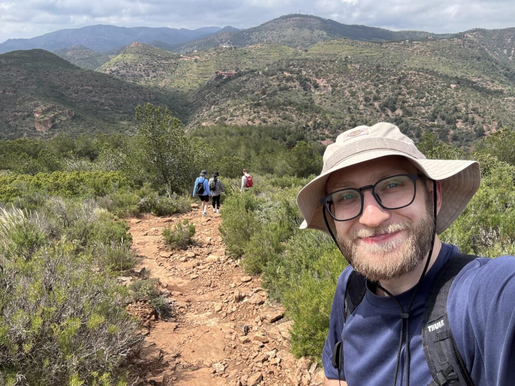 A man wearing glasses and a sun hat smiles at the camera on a rocky hiking trail, as three people walk ahead, framed by green hills and mountains beneath a cloudy sky—capturing the camaraderie of an outdoor interview.