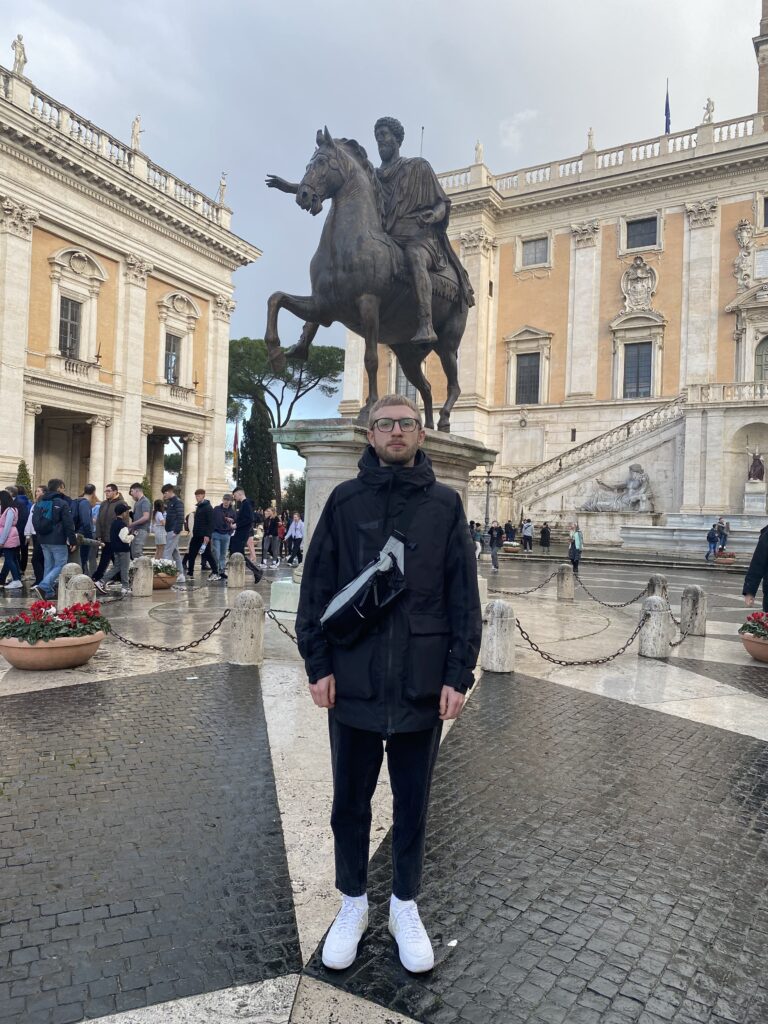 A man in black clothing and white sneakers stands in front of an equestrian statue in a busy square with historic buildings, stone steps, and wet ground—perhaps discussing PPC Reporting during an on-location interview.