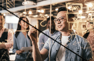 A group of people stand together in an office, watching as a man writes mathematical equations and diagrams on a transparent glass board—collaborating on solutions to prevent harmful ads with Microsoft Advertising. The atmosphere appears collaborative and focused.