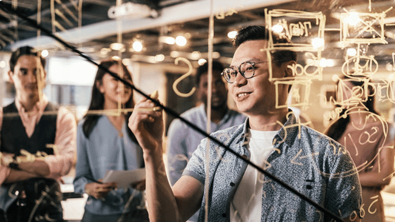 A group of people stand together in an office, watching as a man writes mathematical equations and diagrams on a transparent glass board—collaborating on solutions to prevent harmful ads with Microsoft Advertising. The atmosphere appears collaborative and focused.