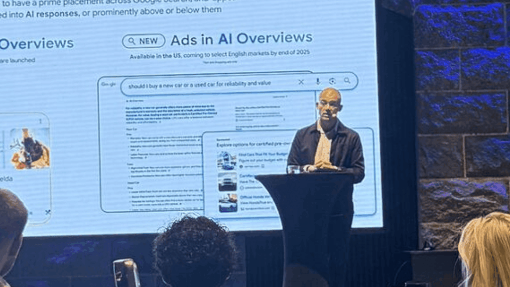 A man stands at a podium speaking to an audience in front of a large screen displaying information about new AI Overviews, Ads, and search features coming in 2025.