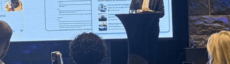 A man stands at a podium speaking to an audience in front of a large screen displaying information about new AI Overviews, Ads, and search features coming in 2025.