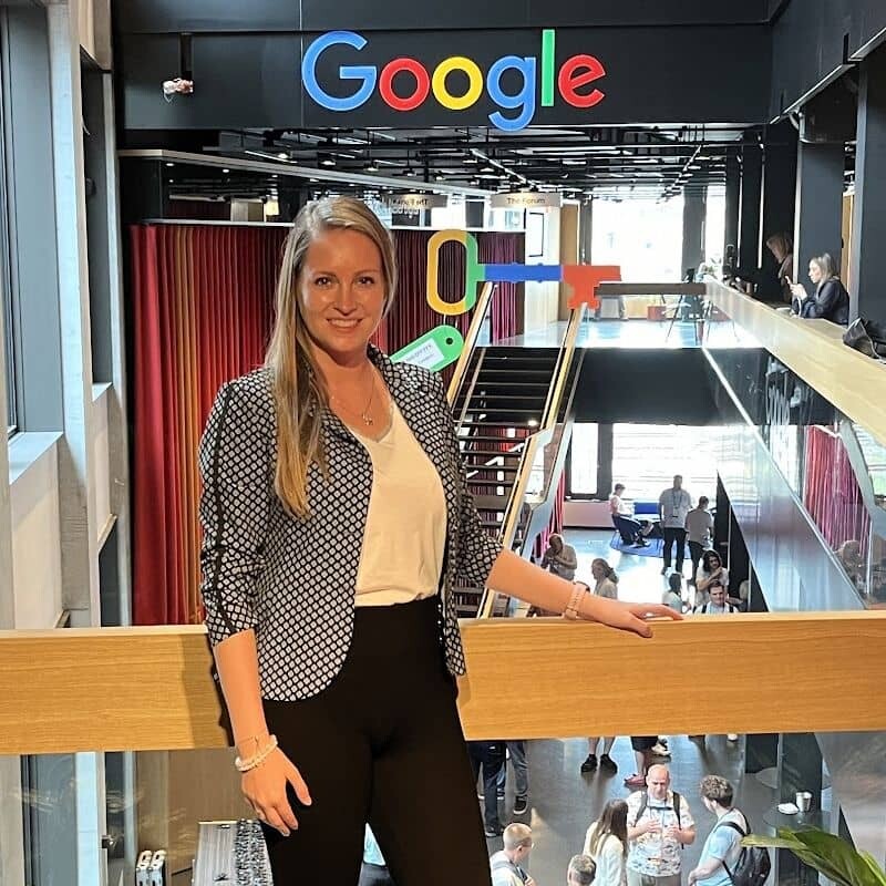 A woman with long blonde hair, wearing a patterned blazer and black pants, stands smiling in front of a railing inside a modern building with a large Google logo—capturing the dynamic world of PPC News unfolding in the open area below.
