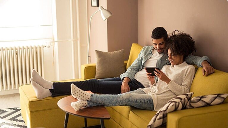 A couple sits closely together on a yellow sofa in a cozy living room, smiling and looking at a tablet device. They appear relaxed, with their legs stretched out and feet resting on a small round table.