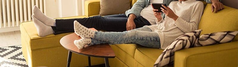 A couple sits closely together on a yellow sofa in a cozy living room, smiling and looking at a tablet device. They appear relaxed, with their legs stretched out and feet resting on a small round table.