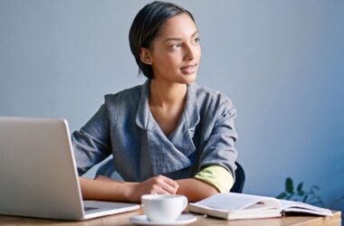 A woman sits at a desk with a laptop, an open notebook, and a cup of coffee. She is looking thoughtfully to the side, wearing a gray jacket, with a calm and focused expression.