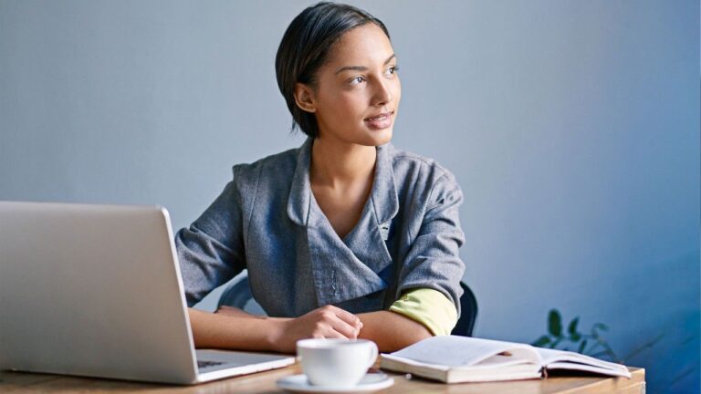 A woman sits at a desk with a laptop, an open notebook, and a cup of coffee. She is looking thoughtfully to the side, wearing a gray jacket, with a calm and focused expression.