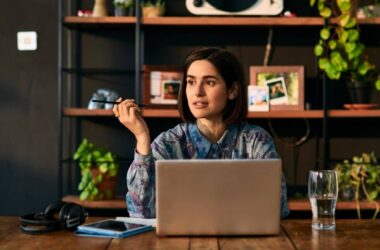 A woman sits at a table with a laptop, holding a pen and looking thoughtful. There are headphones, a notebook, a smartphone, and a glass of water on the table. Behind her are shelves with plants and framed photos.
