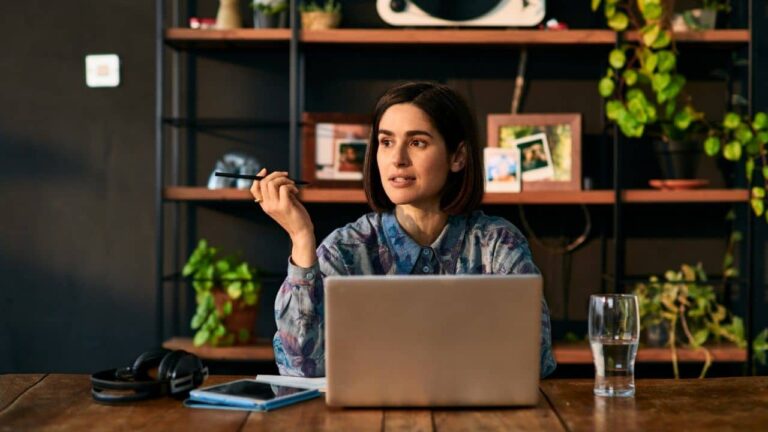 A woman sits at a table with a laptop, holding a pen and looking thoughtful. There are headphones, a notebook, a smartphone, and a glass of water on the table. Behind her are shelves with plants and framed photos.