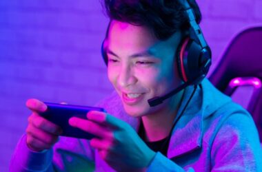 A young person wearing a headset and gray hoodie sits in a gaming chair, smiling and playing a game on a smartphone, illuminated by vibrant purple and blue lighting.