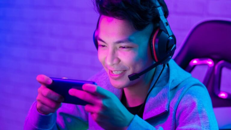 A young person wearing a headset and gray hoodie sits in a gaming chair, smiling and playing a game on a smartphone, illuminated by vibrant purple and blue lighting.