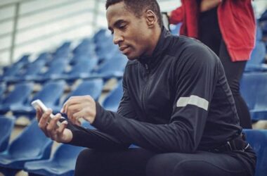 A man in athletic wear sits on blue stadium seats, looking at his phone. Another person in a red jacket stands in the background. The setting appears to be outdoors.