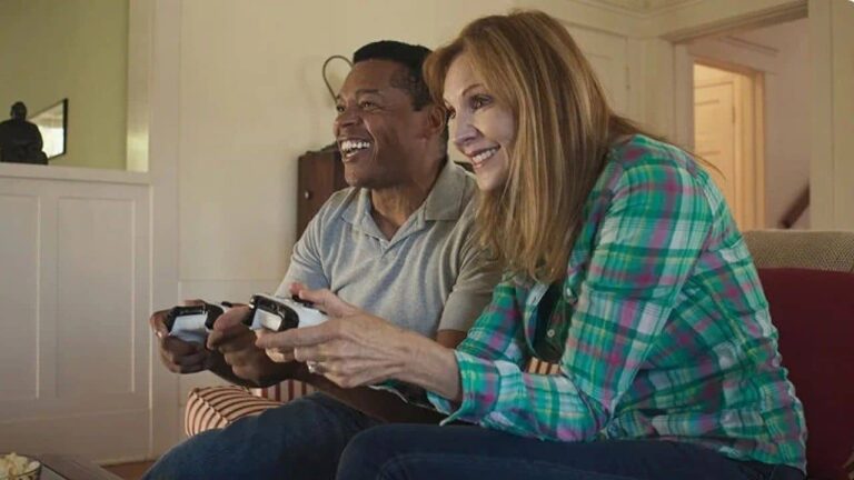 A smiling man and woman sit on a couch playing video games together with controllers in their hands, appearing engaged and happy in a cozy living room.