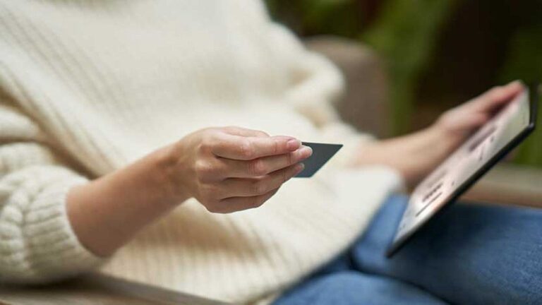 A person in a cream sweater and jeans holds a credit card in one hand and a tablet in the other, suggesting they are shopping or making a payment online. The background is softly blurred.