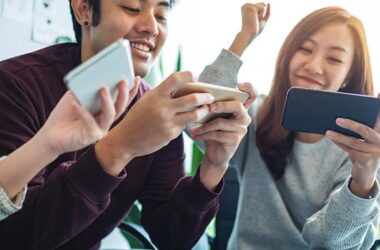 Three young adults smiling and holding smartphones, appearing to play a game or interact joyfully together in a bright indoor setting.