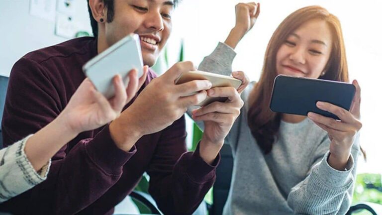 Three young adults smiling and holding smartphones, appearing to play a game or interact joyfully together in a bright indoor setting.