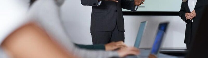 A man in a suit speaks to a group in a meeting room, gesturing with his hands. A woman stands beside him, and blurred attendees are seated at a table with laptops. A data chart is displayed on a screen behind them.