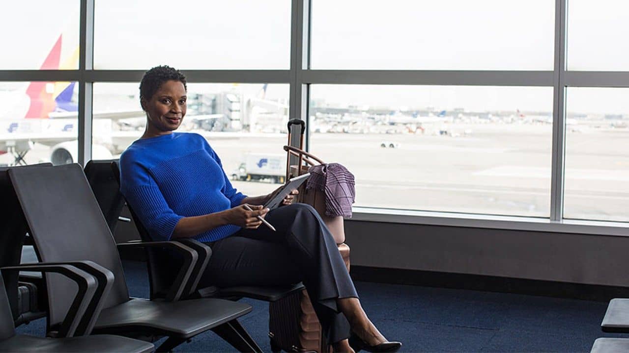 A woman sits in an airport waiting area with a tablet in her hands and a suitcase beside her, looking toward the camera. Large windows behind her show airplanes and the runway outside.